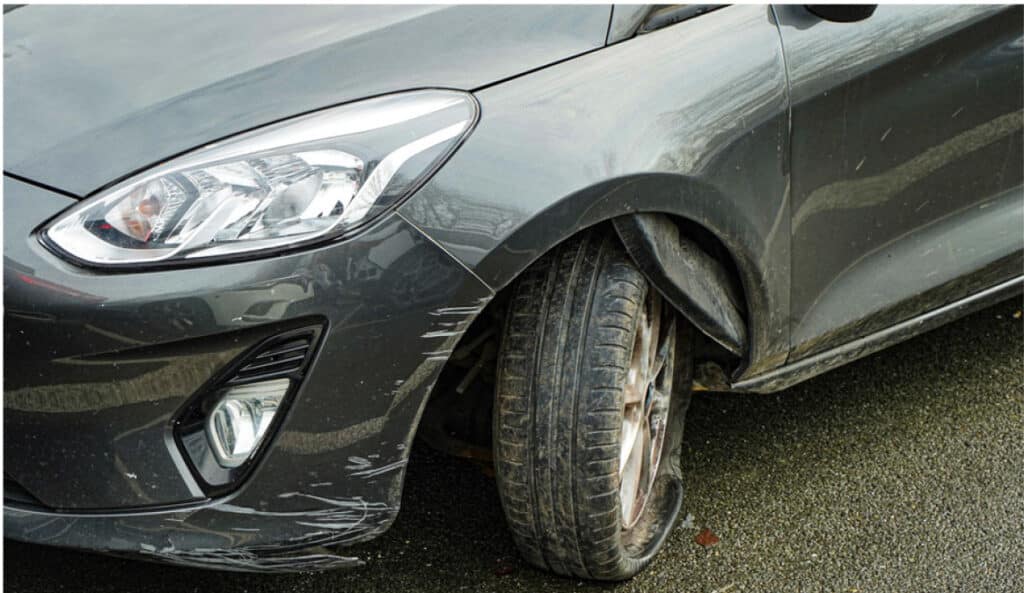 Voiture grise endommagée avec pneu avant gauche crevé et rayures sur le pare-chocs et l'aile.