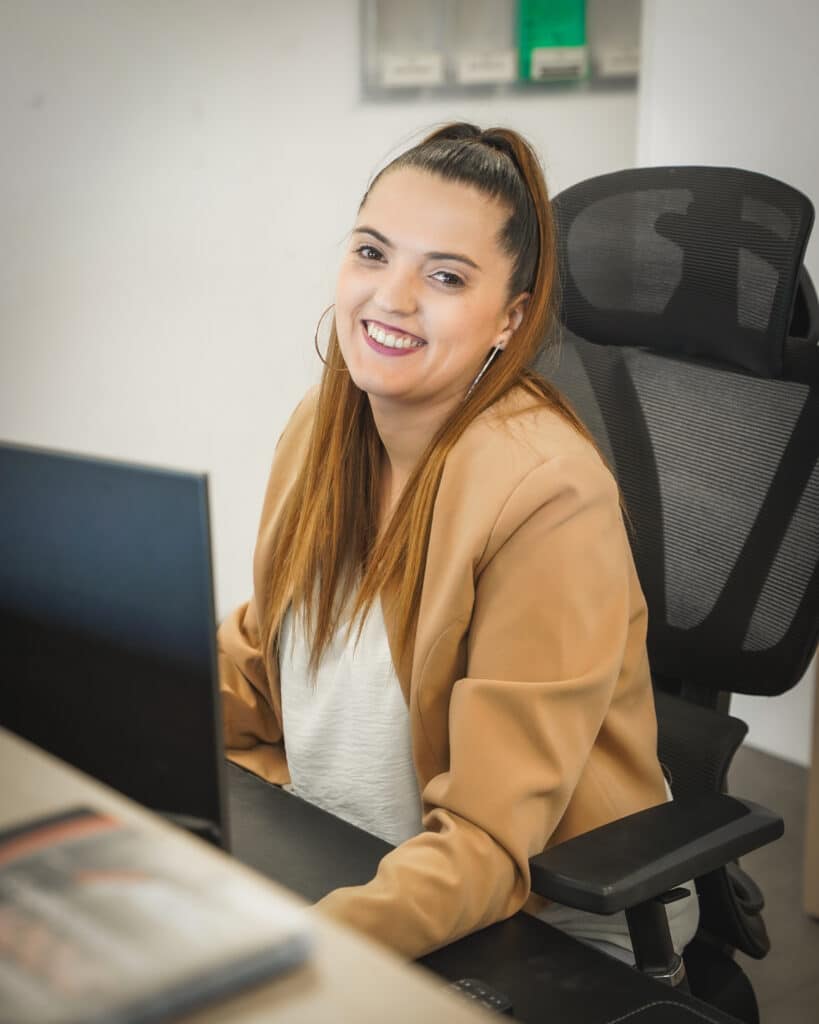 Jeune femme professionnelle souriante en blazer beige, assise à un bureau d'ordinateur dans un environnement de travail moderne.