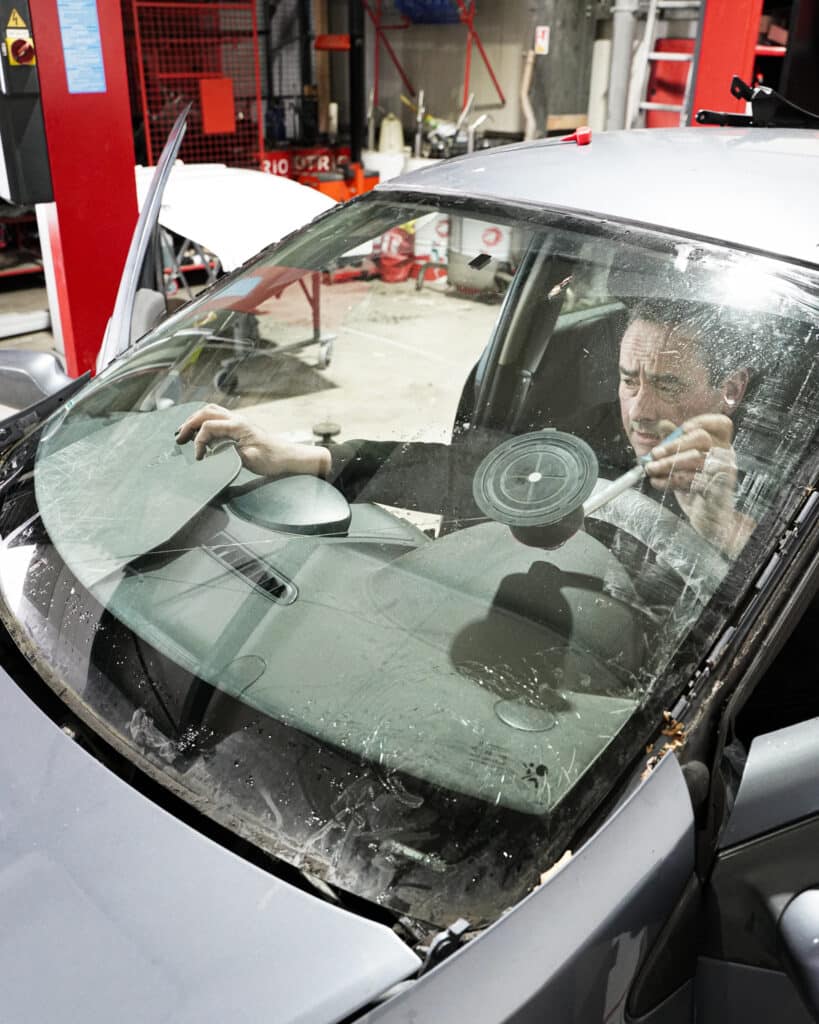 Un mécanicien remplace le pare-brise d'une voiture grise en garage, utilisant des outils dont une ventouse pour le décoller.