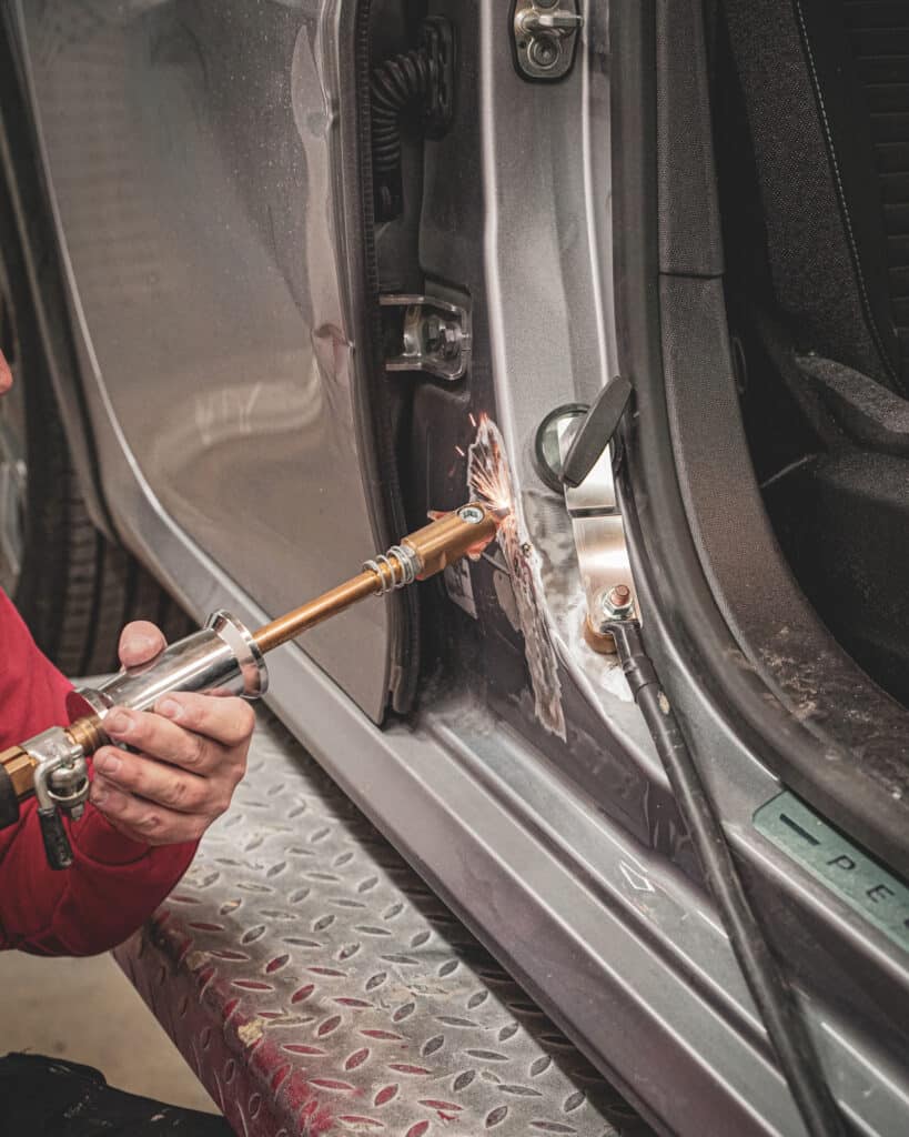 Homme réparant une portière de voiture grise avec un débosseleur à souder, des étincelles volent du métal. Texte "PEUGEOT" visible.