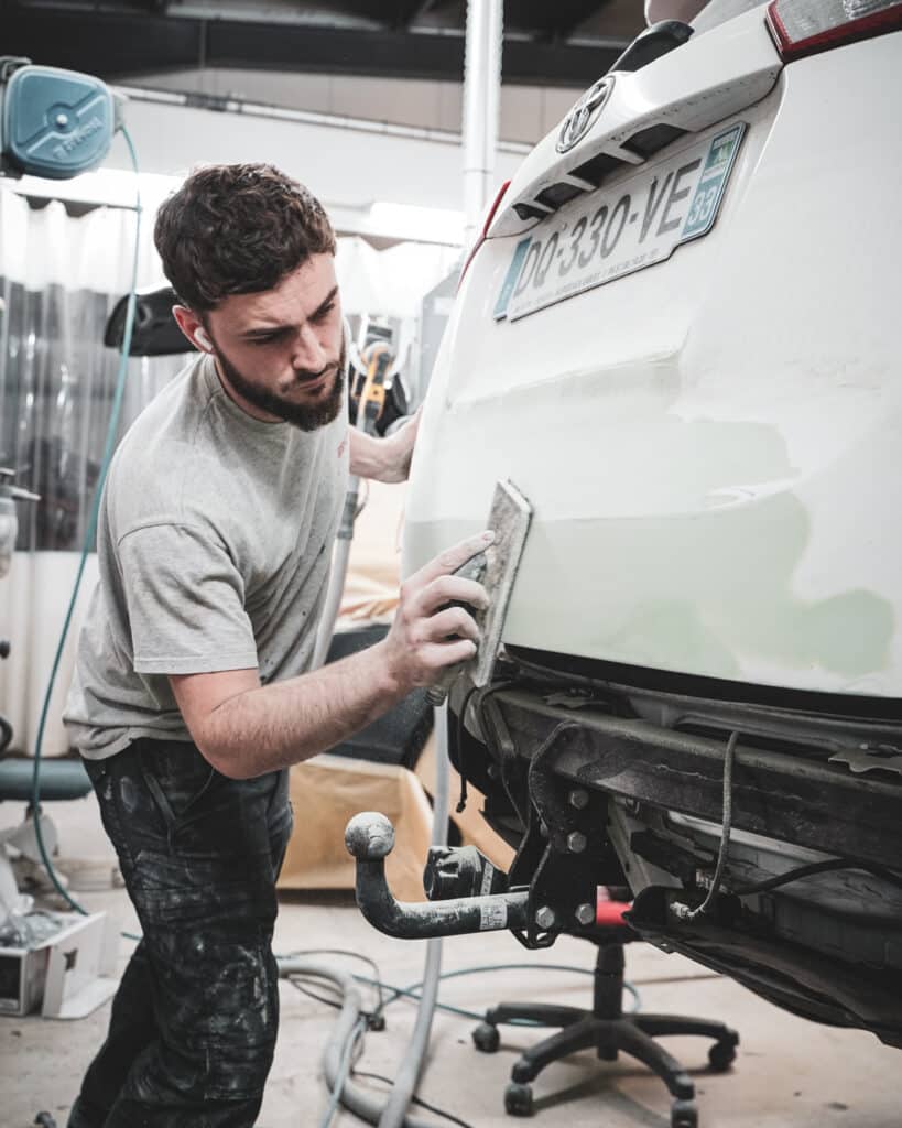 Mécanicien barbu ponçant la carrosserie arrière d'une Toyota blanche en atelier. Plaque d'immatriculation DD-330-VE.