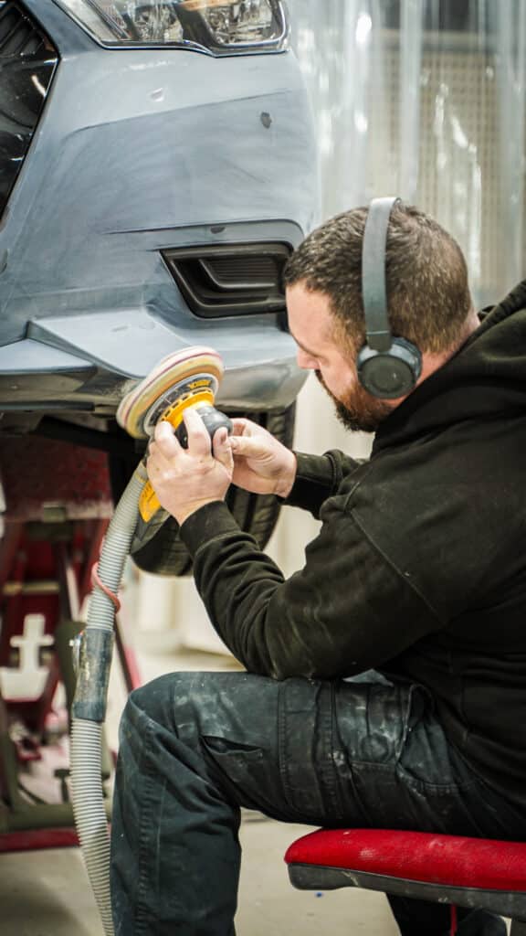 Homme sable le pare-chocs d'une voiture grise en atelier. Il porte des écouteurs et utilise une ponceuse orbitale pour la réparation.